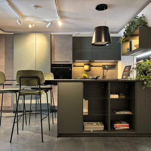 Cucina moderna Life con isola nera, sgabelli verdi, illuminazione led e scaffali aperti con libri e piante decorative.